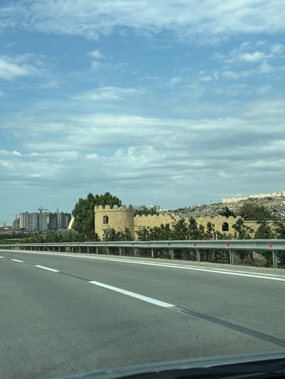 The image captures a scene from a moving vehicle, likely a car, traveling along a highway. The perspective is tilted, suggesting the photo was taken with the device held vertically. The highway itself dominates the right side of the frame, with a gray asphalt surface marked by white lane dividers. A metal guardrail runs along the left edge of the highway, separating it from the landscape beyond.

The landscape is characterized by a dry, hilly terrain. Prominently featured are ancient stone structures, resembling fortifications or towers, built from yellowish bricks. These structures are interspersed with lush green trees. In the distance, a modern cityscape is visible, with numerous high-rise buildings and construction cranes, indicating ongoing development. The sky is a prominent feature, filled with soft, wispy white clouds against a pale blue background.

The time of day appears to be daytime, likely late morning or afternoon, given the brightness and the clear sky with clouds. The weather seems pleasant and fair. The combination of ancient structures and modern construction suggests a location where history meets contemporary urban expansion. The overall impression is one of movement and observation, capturing a blend of historical architecture, burgeoning city life, and natural elements under a wide sky. No people are clearly visible, and no text is discernable in the image. The setting is consistent with the Surakhany Raion in Azerbaijan, known for its historical sites and developing urban areas.