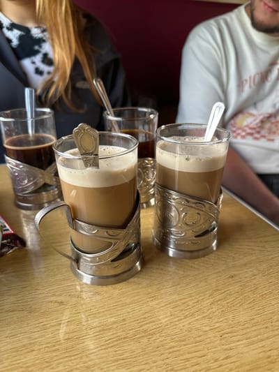 The image shows two glasses of layered coffee drinks served in ornate silver-colored metal holders on a wooden table. Each glass has a spoon inside. Two additional, smaller glasses containing dark liquids are also visible in the background. A person wearing a light-colored patterned shirt is partially visible in the upper left, and another person wearing a black and white patterned garment is partially visible in the lower left. The photo was taken indoors. While the location is indicated as Мажэйкаўскі сельскі Савет, Belarus, there are no visual cues in the image to confirm this specific location within the city.