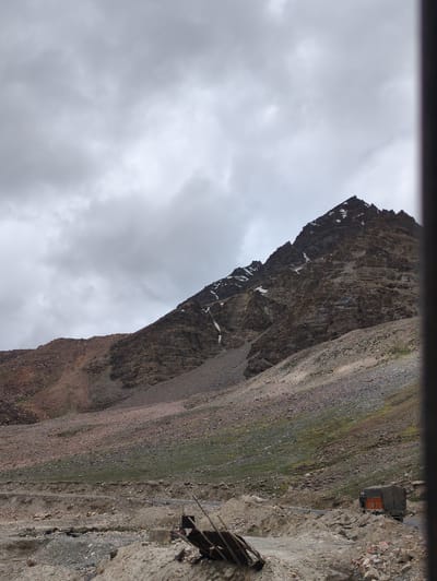 The image depicts a stark mountain landscape under a cloudy sky. A steep, rocky mountain dominates the upper right portion of the frame, with patches of snow visible on its slopes. The lower part of the image shows a rugged, gravelly terrain with a road winding through it. A truck with a covered cargo is parked on the side of the road, partially obscured by debris and construction materials. In the foreground, there are piles of rocks and dirt, suggesting ongoing road work or excavation. The environment appears desolate and remote, characteristic of a high-altitude mountainous region. The scene is devoid of any visible human figures. The location is identified as being in the vicinity of Kelangsarai, India.
