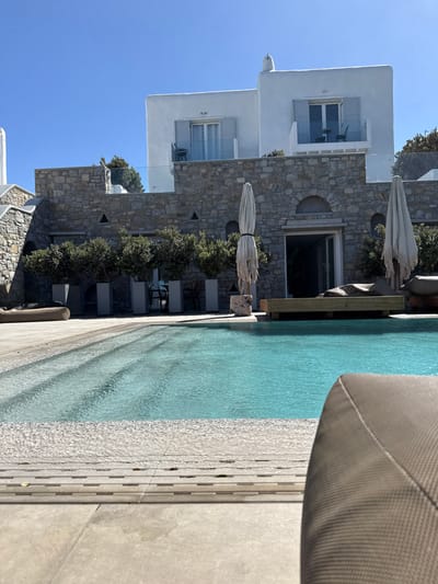 The image displays an outdoor swimming pool area at a property in Mykonos, Greece, bathed in bright sunlight under a clear blue sky. A turquoise swimming pool with visible steps on the left occupies the foreground and mid-ground. The pool is bordered by light-colored paving and wooden decking. A wooden platform lounger with dark cushions is positioned partially submerged in the pool on the right, and a large dark fabric lounger rests on the deck to the far left. The photograph is taken from the low perspective of a dark, textured lounger, a portion of which is visible in the lower right foreground.
In the background, a multi-level building complex typical of Cycladic architecture stands. The lower level is constructed from rough-hewn stone masonry, featuring a central arched entrance with an open dark sliding door. A row of green shrubs in grey planters lines this stone facade. Above this base, two distinct white stucco-finished structures rise. Each features a balcony with light grey louvered shutters flanking a glass sliding door, and clear glass railings. A dark chair is present on the left balcony, and two dark chairs with a small table are on the right balcony. Two large, folded, weathered light-colored beach umbrellas are situated on the pool deck. No individuals are visible.