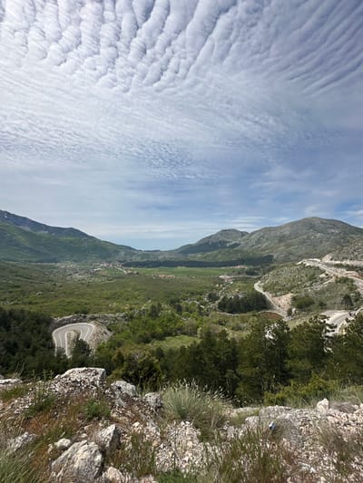 A landscape photograph depicts a mountainous region in Bukovica, Montenegro. The sky, filled with striated clouds, takes up approximately half of the left side of the image. A steep, rocky mountain dominates the right side, covered in sparse vegetation and glimpses of a winding road. Below the mountain is a green valley with scattered vegetation and a few visible buildings. The viewpoint is from a rocky outcrop with small shrubs and grasses.