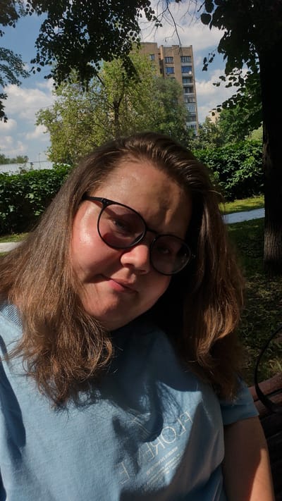 A young woman with brown hair and glasses is taking a selfie outdoors. She is wearing a light blue t-shirt. The background shows trees, a building, and a cloudy blue sky. The location is identified as Павлово-2 Кп, Russia.