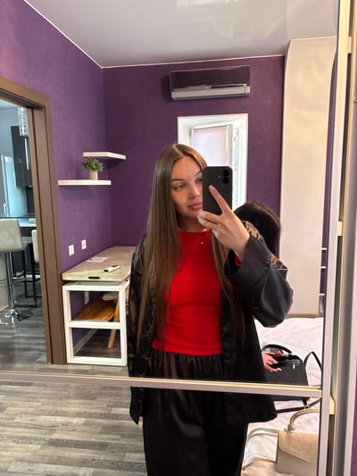 A young woman with long brown hair is taking a selfie in a room with purple walls, reflected in a large mirror. She is wearing a red top and a black open jacket over it, along with black pants. In the mirror's reflection, a shelf with a potted plant is visible on the purple wall, as well as an air conditioner unit. To the left of the woman, a doorway opens into another part of the room or an adjacent room, showing a kitchen counter with stools. Behind her, a bed is partially visible with several bags on it. The floor appears to be tiled with a wood-grain pattern. The scene is indoors, possibly an apartment or hotel room. The location is identified as Voronovka, Russia.