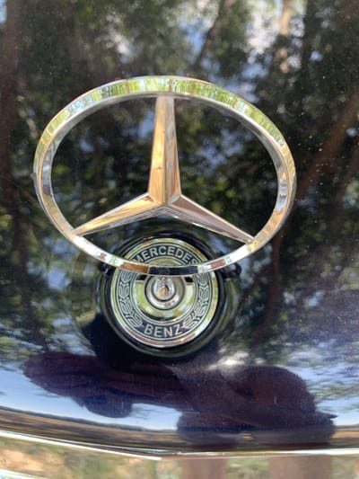 The image is a close-up shot of a Mercedes-Benz logo on a dark-colored car. The logo, a three-pointed star enclosed in a circle, is made of polished metal and reflects the surrounding greenery and sky. Below the star emblem, a circular badge with the text "Mercedes-Benz" is visible. The car's body appears to be a deep, glossy blue or black, with reflections suggesting an outdoor environment with trees and sunlight. The bottom of the image shows a chrome trim piece, likely part of the car's bumper or hood. The scene is set in Indiya, Moldova.