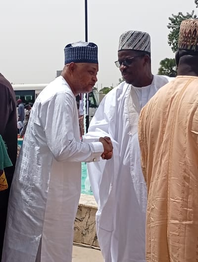 This bright outdoor image, set in Unguwan Sanusi, Nigeria, features two men in traditional attire shaking hands in the foreground, suggesting a greeting or formal interaction. The man on the left wears a white, long-sleeved tunic with a subtle checkered pattern and a dark blue and white embroidered cap. He faces the man on the right, whose hand he holds.

The man on the right is also dressed in a white, long-sleeved tunic and a patterned cap featuring black, white, and possibly brown geometric designs. He wears dark sunglasses and is looking towards the man on the left. To the far right, the back of a third man is visible, wearing a light brown or tan traditional garment with gold embroidery and a patterned cap similar to the man in white.

The scene is brightly lit by the sun, indicating midday or early afternoon, with clear skies. In the mid-ground, behind the handshake, there is a body of water, likely a fountain or a pool, with a decorative stone edge. A light-colored bus or van is partially visible in the left background, while distant trees and foliage, along with a tall dark pole, are seen on the right. The overall impression is a cordial gathering or event in an open, sunny outdoor space. No text is visible in the image.