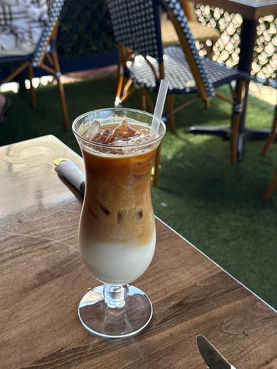 The image shows a cocktail glass filled with an iced coffee drink, positioned on a wooden table outdoors. A white straw is in the drink, which appears to be layered with ice, coffee, and milk or cream. A knife is visible on the table next to the glass. In the background, there are chairs and a green grassy area. The location is likely in Chula Vista, United States of America.