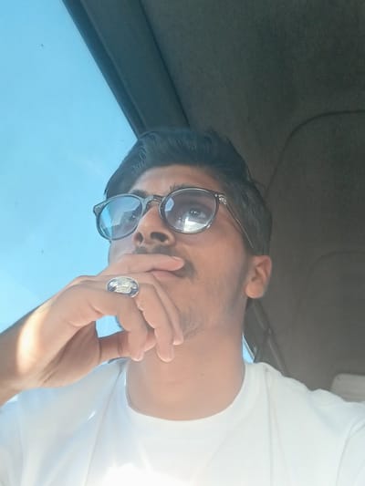 The image is a close-up, low-angle shot of a person wearing glasses and a white t-shirt, indoors, possibly in a vehicle given the dark interior visible at the top and side. The person has dark hair and is looking upwards and to the left. Their right hand is raised, with fingers touching their mouth and chin, and they are wearing a ring with a blue stone and an emblem on their index finger. The background consists of a bright blue sky visible through a window, and a dark, possibly textured surface. The lighting suggests daytime. The location of Aspley Guise, United Kingdom, is not discernible from the visual information provided in the image.
