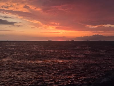 The image shows a sunset over the sea, with the sky painted in hues of orange, pink, and purple. The water is dark and choppy, reflecting the colors of the sky. In the distance, silhouetted against the horizon, are several structures that appear to be stilt houses or small piers. One structure on the left has a distinct roofline, while others are simpler in design. There are also faint outlines of ships or boats further out at sea. The overall impression is one of a peaceful and serene evening by the water.