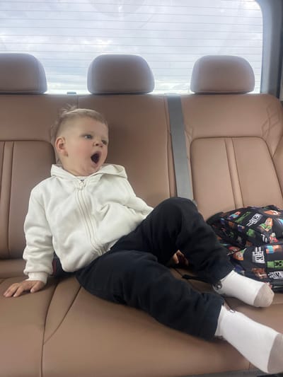 A young child with blonde hair is sitting in the back seat of a vehicle. The child is wearing a white zip-up hoodie and black pants. They are yawning widely, with their mouth open and eyes partially closed. A backpack with a Minecraft pattern is visible next to the child. The car's interior is upholstered in brown leather, and headrests are visible in the background, along with a portion of the rear window showing a cloudy sky. This scene is not located in Dybshche, Ukraine.