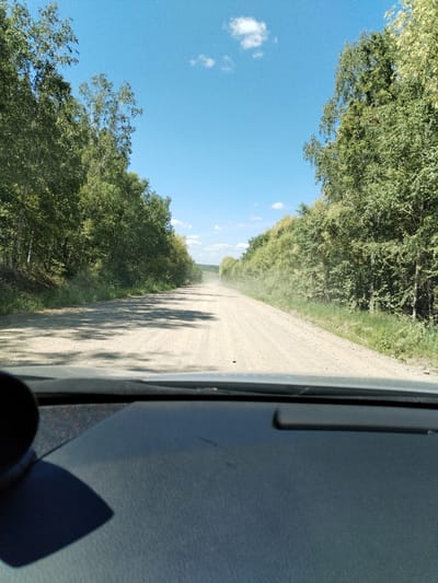 The image shows a dirt road curving into the distance, flanked by dense green trees on either side. The road is dusty, with a visible cloud of dust trailing behind a vehicle that is not in frame. The sky is a clear blue with a few small white clouds. The foreground shows the dashboard of a car, suggesting the photo was taken from inside the vehicle. The location is given as Antonovka, Russia.