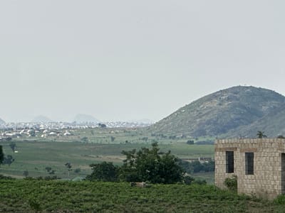 The image depicts a rural landscape with a partially constructed building in the foreground. The building, made of light-colored bricks, has two dark, rectangular window openings visible. Behind the building, a vast expanse of green fields stretches towards a distant town. The town, characterized by numerous white-roofed structures, is nestled at the base of a large, green hill. The hill, prominent in the mid-ground, features a rocky, uneven surface and is partially obscured by a hazy sky. The overall impression is one of ongoing development in a semi-arid, hilly region, likely in Gidan Waziri, Nigeria, given the context. The sky is overcast, contributing to the muted lighting of the scene. No people are visible in the frame.