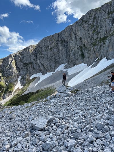 In Pašina Voda, Montenegro, two individuals are depicted in a mountainous, rocky terrain. One person, wearing a hat and a dark shirt, stands on a large rock, holding their hand to their forehead as if shielding their eyes from the sun. The other person, in the foreground and to the right, is wearing a black t-shirt, white shorts, and a black hat, and is holding a smartphone. The environment is characterized by a steep, rocky mountainside with patches of snow and sparse green vegetation. The sky above is blue with scattered white clouds. The ground is covered in loose grey rocks of varying sizes.