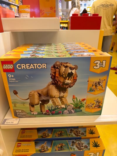 A LEGO Creator 31386 Majestic Lion building set is displayed on a store shelf in Mission Viejo, United States of America. The set is a large cardboard box with the LEGO logo and "Creator" branding prominent. The box features an image of a large lion constructed from LEGO bricks, with smaller images of alternative builds, including a lioness and cubs, and a smaller lion. The price tag for the set is $69.99. Below the main display, there are stacked boxes of another LEGO Creator set, 31388 "Famille de koalas" or "Koala Family". In the background, people can be seen browsing in the store, with yellow and red LEGO-themed displays visible.
