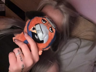 The image features a close-up of a hand holding an orange, plush toy of a character resembling Naruto. The hand has a tattoo on the fingers reading "SOON AFTER" and is adorned with rings. The toy is held against a background of gray-blonde hair, suggesting the person is lying down. Parts of the background are blurred. There is no indication of the location being Obruchishte, Bulgaria, in the image.