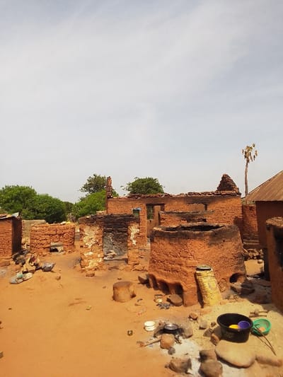 The image shows the aftermath of a fire in a village in Kwal, Nigeria, at coordinates 9.803 latitude and 8.646 longitude. The scene is a ruined mud-brick structure with charred walls and a collapsed roof. The surrounding ground is covered in red sand, and the remnants of burnt debris are scattered about. Several cooking pots and utensils are visible, as well as a sack of grain and a plastic bucket. There are no people visible in the image. The sky is a light blue with some white clouds. A single palm tree stands in the background.