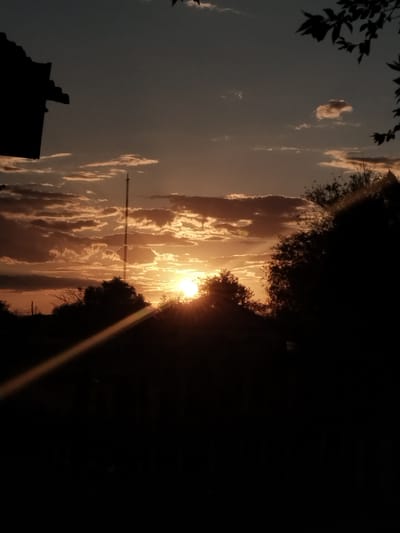 The image depicts a sunset in the city of Ақадыр, Kazakhstan. Silhouetted against the vibrant, orange-hued sky are several elements, including a tall, slender radio tower with a lattice structure, trees with dark foliage, and what appears to be part of a building or structure on the left side of the frame. The sun is low on the horizon, casting a bright, radiant glow that peeks through the clouds and trees, creating a lens flare effect with a distinct diagonal streak of light across the lower portion of the image. The sky is a gradient of deep blue at the top transitioning to warm oranges and yellows near the horizon, with scattered clouds catching the sunlight. The overall scene is one of a rural or semi-rural landscape at dusk.