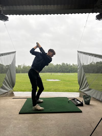 A young man, wearing black pants and a dark blue jacket, is swinging a golf club from a covered driving range. The driving range is located in South Ribble, United Kingdom. The range is open to the elements, with only a partial roof and mesh netting on the sides to contain errant golf balls. The man is facing the right side of the frame with his back to the camera, and he appears to be in the middle of his backswing, as the club has reached behind his head. The range is lined with trees in the distance. There are several golf balls on the ground. The sky is overcast, with a few scattered clouds. The ground is wet from recent rain. There is a tee in the ground, and a bucket full of golf balls next to the tee. It is raining lightly.
