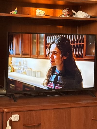 A television screen displays a scene of a young woman with long, dark, curly hair, looking distressed. She is wearing a striped sweater with blue, red, and white stripes. The background on the screen appears to be a kitchen with wooden cabinets and white countertops.
The television is placed on a wooden cabinet. On the shelf above the television, several small decorative items are visible, including a small figurine of a duck or bird, a small rectangular object with a picture of a building, and what appears to be a white folded cloth or piece of paper.
The cabinet itself is made of wood and has a reddish-brown finish. A white childproof lock is attached to the front of one of the drawers. The overall environment suggests a domestic setting. The image is taken from an angle that captures the television screen and the furniture it rests on, with the surrounding shelving.