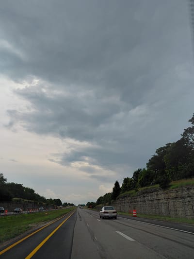The image shows a view from a vehicle driving on a highway in Midway, United States. The scene is dominated by a dramatic sky filled with dark, heavy clouds, suggesting impending rain or stormy weather. The sun is not directly visible, but there are brighter patches in the sky, indicating it might be late afternoon or early evening.

On the right side of the frame, the highway stretches ahead, marked with yellow and white lines. A white car is visible in the right lane, further down the road. The road is bordered by a grassy median and a steep, rocky cliff face covered with trees. Orange traffic cones are placed along the side of the road, indicating potential construction or a hazard. Several other vehicles, including a truck, are visible in the distance.

The left side of the image is primarily occupied by the sky and trees along the top edge. The perspective suggests the camera is tilted, giving a wide-angle view of the highway and its surroundings. The overall mood is somewhat somber due to the dark clouds, but the presence of the highway and vehicles suggests ongoing travel and activity. No visible text is present in the image.
