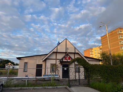 The image shows a modest, light-colored building with a pitched roof and a cross on top, resembling a small church or chapel. The facade has decorative wooden beams and an arched entrance. Red Cyrillic letters are visible above the entrance. The building is surrounded by a metal fence. To the right, a modern brick apartment building stands tall. A street lamp is positioned near the apartment building. Green foliage, including a thick vine covering part of the fence, adds to the scene. The sky is filled with scattered clouds under a blue sky, suggesting it is daytime, possibly late afternoon due to the warm light on the apartment building. A blue bench is visible in front of the building. The overall impression is of a mixed urban and semi-rural setting. The building is located in Balabanovo, Russia.