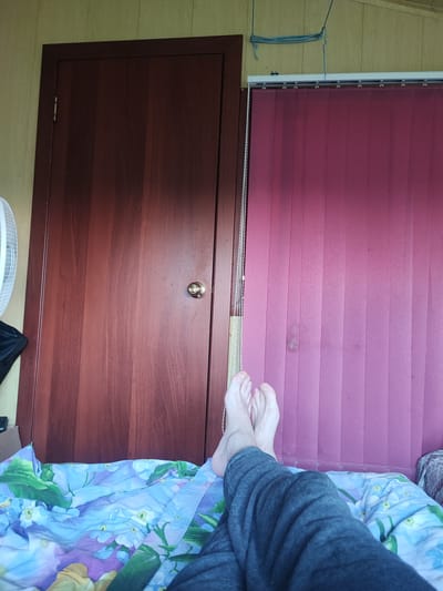 The image shows a person's legs and feet resting on a bed covered with a floral patterned duvet. The person is wearing grey sweatpants. Behind the bed, there is a closed wooden door with a golden doorknob. To the right of the door, there are vertical blinds in a dark pink color. A white fan is partially visible on the left side of the image. The setting appears to be a bedroom. The lighting suggests it is daytime. There is no visible text in the image.