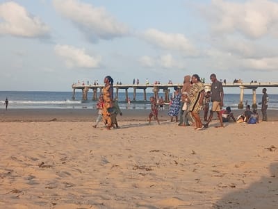 This image depicts a busy beach scene in Shella ward, Kenya. The foreground consists of light brown sand with scattered dark organic debris. In the midground, a significant number of individuals are present on the beach. A central group of approximately eight people is walking from left to right. This group includes adult females, one wearing a yellow and black patterned dress and holding an infant, and another wearing a brown hijab. A male in a dark t-shirt and light shorts walks barefoot beside them. To the right of this group, several individuals are seated or reclining directly on the sand, some with visible personal belongings. Further right, a lone male in a dark t-shirt and shorts stands near the water's edge. On the far left, a solitary figure stands partially submerged in the ocean. The background features the ocean with small waves breaking near the shore, and a long concrete pier extending into the water. Numerous individuals, estimated at 20-30, are visible walking, standing, and congregating along the entire length of the pier. The sky is light blue with scattered white cumulus clouds, indicating an outdoor, possibly late afternoon, setting.
