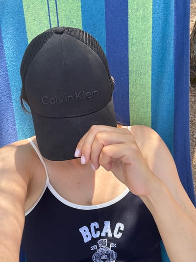 A woman wearing a black Calvin Klein baseball cap and a black tank top with "BCAC" printed on it is lying down on a striped blanket. Her hand is covering her face, and her nails are painted a light pink color. The blanket has alternating stripes of blue and green. The scene appears to be outdoors, possibly in a park or by a body of water, given the sunlight and shadow play on her skin. There is no indication of the location being in Антонов ауылдық округі, Kazakhstan.