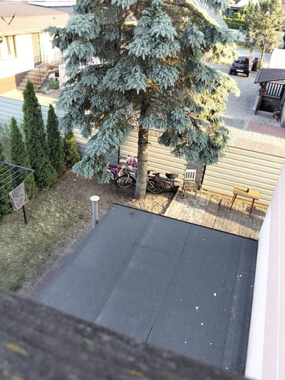This media file depicts a residential backyard in Brodnica, Poland. The scene is viewed from an elevated perspective, likely a balcony or window, looking down into a yard with a large blue spruce tree prominently in the foreground. The yard is partially paved with interlocking stones and partially covered in dry grass and soil.
Visible objects include two bicycles leaning against the trunk of the spruce tree, one of which has a purple bag attached to its handlebars. Nearby, there is a weathered wooden chair and a small wooden table with a small object on top. A clothesline with a towel hanging on it stands in the left side of the yard, near a row of tall, conical evergreen trees. A light-colored cylindrical pipe with a cap stands in the middle of the yard. Beyond the yard, a light gray corrugated metal fence separates it from a paved driveway where a dark-colored car is parked. A small, covered structure with railings and potted plants is also visible in the distance, along with parts of a house and roof.
The lighting suggests it is late afternoon or early evening, with the sun casting long shadows. The weather appears to be fair, and the overall impression is one of a quiet, everyday scene in a suburban or rural setting. No people are visible in the frame. There is no visible text in the image.