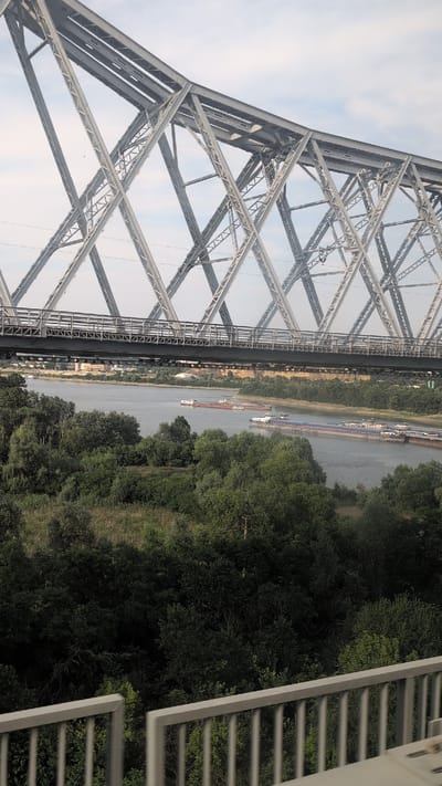 This is a photograph of the Borcea River in Borcea, Romania, as viewed from a bridge. A large, steel truss bridge spans across the river. Several barges are visible on the water, appearing to be in transit. The riverbanks are lined with lush green trees. In the background, a town is visible on the opposite bank, with buildings and structures suggesting a settled area. The sky is partly cloudy. The foreground shows a portion of the railing of the bridge from which the photograph was taken.