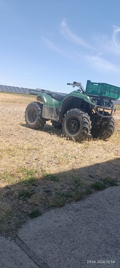 In Berezanka, Ukraine, a green ATV is parked on dry, grassy land with some scattered rocks. The ATV has large, rugged tires and a green plastic crate is attached to a rack on the back. In the background, a solar farm with rows of solar panels stretches across the horizon under a clear blue sky with wispy clouds. The foreground shows a section of concrete pavement with some sparse green vegetation growing at its edge. The image appears to be taken on a sunny day.