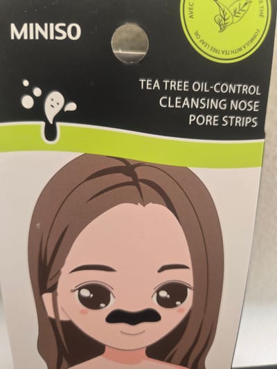The image shows the front of a package of MINISO "TEA TREE OIL-CONTROL CLEANSING NOSE PORE STRIPS". The packaging features a cartoon illustration of a young woman with brown hair, large eyes, and a black nose strip. The text "MINISO" is printed in white on a black background at the top of the package, along with a circular emblem containing text and a leaf design. Below this, the product name is displayed in white text. The bottom portion of the image reveals the upper part of the cartoon character's face and shoulders. The packaging appears to be made of glossy material and is likely a retail product. There is no discernible indication of a location within the city of Deira, United Arab Emirates, as the image is a close-up of product packaging.