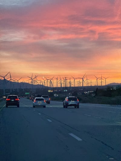 The image captures a scene from a highway in the city of Desert Hot Springs, United States. The view is from the perspective of someone driving on the road, with several cars visible ahead. In the background, there is a field of wind turbines, and mountains on the horizon. The sky is colored with orange and pink hues suggesting a sunrise or sunset.