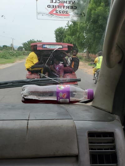 A partially opened bottle of purple liquid rests on the dashboard of a vehicle. In the background, a red vehicle is being used to transport several people. Some individuals are seated in the back of the red vehicle, while one person is riding a motorcycle in the distance. The scene appears to be on a paved road, with vegetation and trees visible on either side. The sky is overcast. A sticker is visible on the windshield of the vehicle in the foreground, with text that is partially obscured and appears to be in a foreign language. The overall impression is a candid snapshot of a journey in Kwadon, Nigeria.