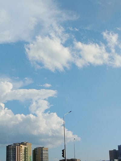 The image shows a clear blue sky with scattered white clouds. In the foreground, there are several apartment buildings of varying heights and colors. One prominent building on the left is light brown with some greenish sections. To its right, another building is a light beige color. In the middle ground, a tall, slender lamppost with a curved top is visible against the sky. Further in the distance, another, shorter lamppost is also present. The scene appears to be a residential area with urban infrastructure under a bright sky.