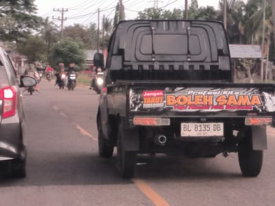 The image shows the rear of a black pickup truck on a road in Damatutong, Indonesia. The license plate reads "BL 8135 DB" and has "10-29" below. The truck's tailgate has a sign with the words "Jangan Takut Boleh Sama." Several motorcycles are also visible on the road in front of the truck. The environment appears to be a residential area, with trees and houses visible in the background. Electrical poles with wires are present on the side of the road. A portion of a gray car is visible on the left side of the image. A yellow line divides the road.