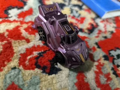 The image displays a metallic purple toy vehicle, a stylized race car model, positioned on a patterned rug in the city of Pakhtakor, Uzbekistan. The toy car features black wheels, a prominent rear spoiler with a gold-colored winged emblem, the numeral "6" on its roof, and gold chevron-like markings on the front hood. The rug beneath the car exhibits an abstract design comprising irregular shapes in red, beige, black, and light green tones. In the upper right portion of the frame, a partially visible, out-of-focus blue rectangular object is present. No individuals are depicted, and no actions or interactions are occurring within the scene.