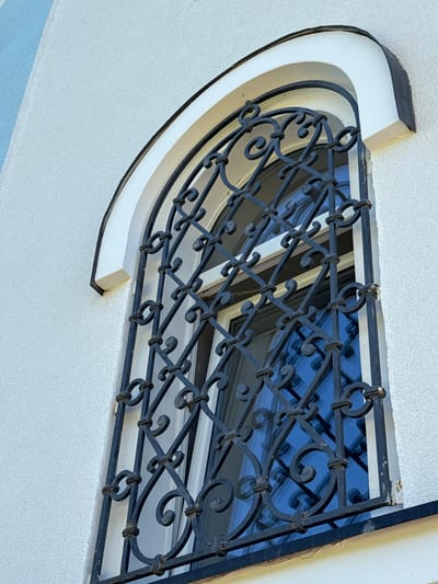 This is a close-up, eye-level outdoor shot of a decorative wrought-iron window grill on a white stucco building. The grill features a diamond pattern with ornate scrollwork at the intersections. The window itself has a blue reflection, suggesting it's either tinted or reflecting the sky. Above the window, a curved architectural element, painted white with black trim, frames the top of the opening. Part of a light blue wall is visible to the left of the window. The building appears to be in St. Ozerki, Russia, though no specific landmarks or people are identifiable. The image focuses solely on the architectural details of the window and its surroundings.