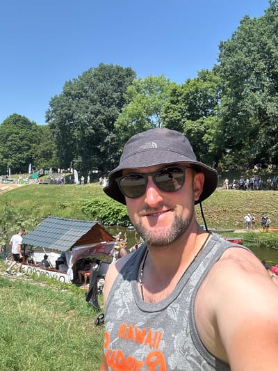 The image depicts a man in a bucket hat and sunglasses taking a selfie outdoors on a sunny day. He is wearing a grey tank top with an orange design. In the background, there is a body of water, a small structure on the water with a roof, grass, trees, and a crowd of people on a grassy slope. The scene appears to be an outdoor event or gathering by a river. The specific location in Racibórz, Poland, is likely the Oder River during a summer event, possibly a regatta or festival, given the presence of boats, a crowd, and recreational activities.