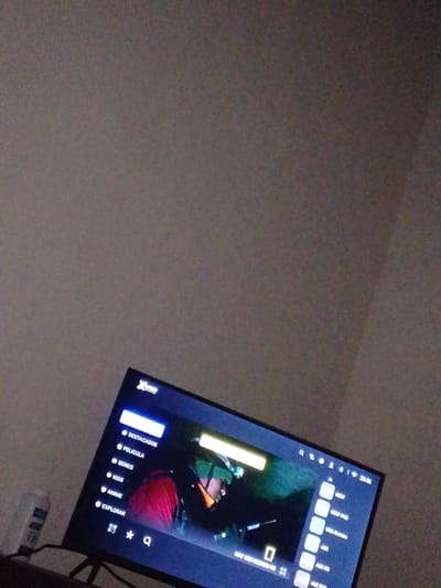A dimly lit, indoor scene captures a television screen displaying a streaming service interface. The screen, positioned on a stand, shows a menu on the left with options like "DESTACADOS" (Featured), "PELICULA" (Movie), "SERIES", "KIDS", "ANIME", and "EXPLORAR" (Explore). The right side of the screen features a video playing, with a person wearing a red jacket visible. The interface includes icons for search, user profile, and settings, along with a timestamp reading "23:56". To the left of the television, a white bottle with a label is partially visible. The background is a textured, light-colored wall, suggesting the setting is a residential interior. The overall lighting is low, with the primary illumination emanating from the television screen. The image is grainy, indicating a potential issue with low light conditions or camera quality. The location is identified as Atamo Sur, Venezuela.
