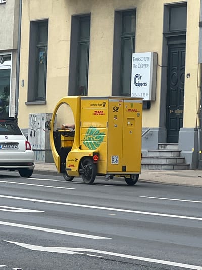 This is a photograph of a yellow DHL delivery vehicle, possibly a cargo bike or a small electric van, parked on a street in Aachen, Germany. The vehicle is bright yellow with "DHL" branding and a leaf logo visible on its side. A driver is partially visible inside the vehicle's open compartment. The scene appears to be set during the daytime. In the background, a building with a light-colored facade features a sign for "FAHRSCHULE DR. CUPPERS" (Driving School Dr. Cuppers) with a phone number. The street has clear lane markings, indicating it's a road for vehicular traffic. A white car with a German license plate "AC AK 1912" is parked behind the DHL vehicle. The overall impression is a typical urban street scene in Germany, with a focus on a delivery vehicle in operation.
