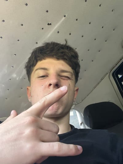 An individual is captured in a close-up, selfie-style photograph, looking directly at the camera with one eye open and the other squinting. Their right hand is raised, with the index finger extended towards the viewer, positioned just in front of their face. The background consists of a textured, light-colored ceiling with numerous small, dark circular indentations, possibly a vehicle's interior. A portion of a dark car seat is visible to the right of the individual. The lighting appears to be ambient, creating some shadows on the person's face and hand. No specific location in Taran, Bulgaria is discernible from the image.