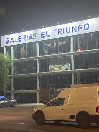 This nighttime image captures the exterior of a multi-story commercial building, identified by a large, illuminated sign reading "GALERIAS EL TRIUNFO" in prominent blue capital letters. The building features extensive glass windows, through which various displays are dimly visible. These displays include what appear to be decorative items, possibly household goods or furniture, and a large, brightly lit circular or arched graphic visible on one of the upper floors.
On the right side, a light-colored delivery van, possibly white or beige, is prominently featured on the adjacent road. The word "ROBUST" is distinctly visible in a vertical orientation on the side of the van. Further in the background, a smaller, light-colored car is also present on the road.
The dark sky confirms it is nighttime, and the building's sign and interior lights provide the primary illumination. Some green foliage is visible at the base of the building. The scene depicts a commercial setting, likely a shopping center or department store, in Puerto Vallarta, Mexico.
