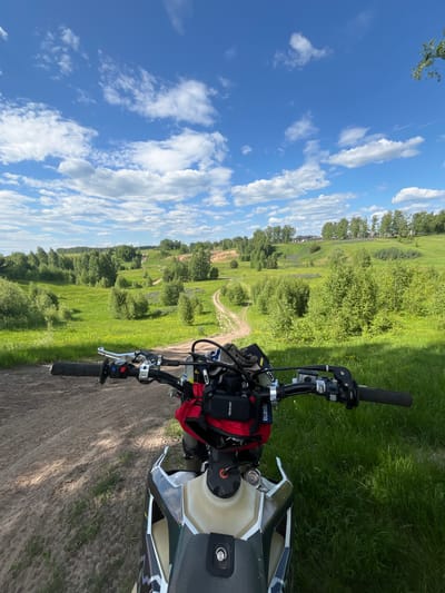 This first-person point-of-view image, captured in a rural area near Новочувашский, Russia, depicts the front view from a stationary off-road motorcycle on a dirt track. In the immediate foreground, the motorcycle's black handlebars are visible, equipped with handgrips, brake and clutch levers, and control buttons (red and blue). A red utility bag is mounted centrally beneath the handlebars, alongside a GPS or communication device bearing "panaride" branding and "husqvarna" on the handlebar pad. The front portion of the motorcycle's fuel tank, in beige and white with dark accents, is also visible.

The scene beyond the motorcycle unfolds into an expansive, hilly green landscape under a bright blue sky dotted with scattered white cumulus clouds. A winding, unpaved dirt path extends directly forward from the motorcycle, disappearing into a valley. The terrain is characterized by rolling hills covered in lush green grass and numerous deciduous trees and bushes of varying sizes. Patches of small purple and yellow wildflowers are discernible in the grass. In the far background, atop a distant hill, a cluster of buildings, possibly residential or farm structures, is visible amidst a line of trees. The lighting suggests a clear, sunny day. No individuals are present in the image.