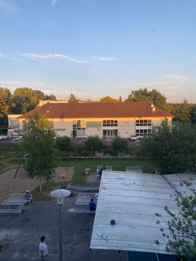 The image captures an outdoor scene in Göttingen, Germany, likely during the late afternoon or early evening, given the warm sunlight illuminating the buildings. In the background, a long, single-story building with a reddish-brown tiled roof is prominent. The building has large windows and signage that reads "ASC SPORTZENTRUM". Cars are parked in front of and beside the building. In the middle ground, there is a playground area with a swing set and a sandpit. Several trees and bushes are scattered throughout the scene, with a larger, leafy tree to the left of the building.

The foreground features an area with picnic tables and benches, some of which are occupied by individuals. A lamppost stands in the center of this area. Further in the foreground, there is a sloped, flat surface covered with light-colored planks, with various objects scattered on it, suggesting it might be a rooftop or an elevated platform. A person in a white shirt and patterned pants is standing and looking to the right in the bottom left corner of the frame. The sky above is a clear blue with some wispy white clouds. The overall impression is a communal space, possibly a park or a recreational area adjacent to a sports center.
