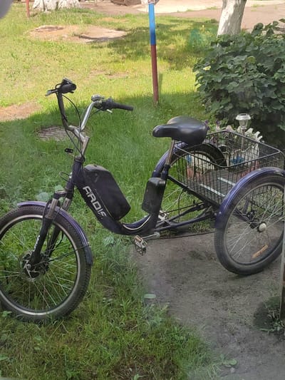 A purple electric tricycle is parked on a dirt path next to a grassy area with some weeds. The tricycle has a basket on the back filled with various items, including what appears to be bottles and papers. The front of the tricycle features a battery pack mounted on the frame, with the word "ARDIS" visible in white lettering. The handlebars have grips, a bell, and a brake lever. The tricycle has three wheels, with the front wheel being larger than the rear wheels. The background includes a bush, a tree trunk, and a pole with blue and red paint. The ground is a mix of dirt and grass. The scene is outdoors during the daytime, with sunlight casting shadows. No people are present in the image. The location is specified as Voskobiinyky, Ukraine.