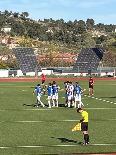 A soccer game is in progress on a green field in Valpaços, Portugal. A team wearing blue and white jerseys is grouped together, while a player in a red jersey lies on the ground nearby. A linesman in a yellow shirt and black shorts stands on the sideline, holding a yellow and orange checkered flag. In the background are two sets of solar panels, a red running track, a view of the city with houses and trees, and a blue sky.