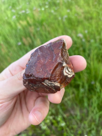 A hand holds a raw, reddish-brown piece of amber against a blurred background of green grass. The amber is irregularly shaped, with a translucent, polished surface that reflects light. Some lighter, yellowish-white inclusions are visible on the upper portion of the amber. The hand appears to be that of an adult, with fingernails clearly visible. The setting suggests an outdoor environment, likely a grassy area. The lighting appears natural, indicating it could be daytime. There is no visible text in the image.