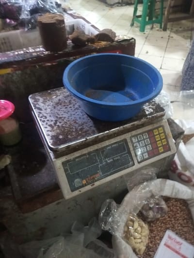 A used digital scale sits on a worn surface, topped with a blue plastic bowl. Next to the scale are plastic bags filled with nuts and grains. Beside the scale, on the same surface, is a chunk of brown sugar. The scale's surface is stained and has visible wear and tear. Other items, possibly food products, are partially visible in the background.