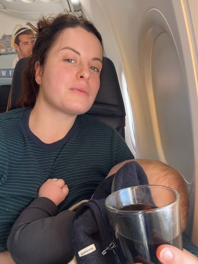The scene depicts the interior of an aircraft cabin, understood to be associated with the city of Knokke-Heist, Belgium. A woman, appearing to be in her late 20s or early 30s, occupies a dark-colored aircraft seat on the left side of the frame, facing the camera with a neutral expression. She has light skin and dark hair pulled back. She wears a dark blue t-shirt with thin green horizontal stripes. An infant is positioned against her chest, partially obscured, with visible light brown hair on its head and a dark-colored zipped garment, possibly fleece, with a beige lining and a white "hessnatur" label. The infant's small, light-skinned hand is visible near the woman's chest. The woman's right hand holds a clear glass containing a dark brown liquid, positioned in the lower right foreground. An airplane window with light visible through it is to the right of the subjects. Behind the woman's head, an advertisement or decorative poster is visible on a cabin partition, featuring a man in a fedora hat with a light-colored background and partial text "W F".