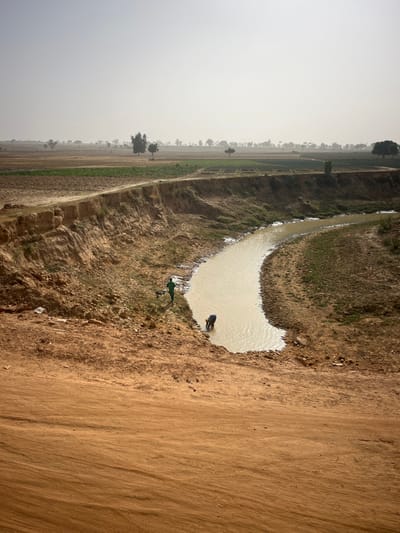 A muddy landscape features a curved waterway filled with tan-colored water. A dark figure stands at the water's edge, and a green object lies nearby on the bank. The right side of the image shows a dirt path. On the left, cultivated fields stretch into the distance under a hazy sky. The horizon is obscured by the dense atmosphere.